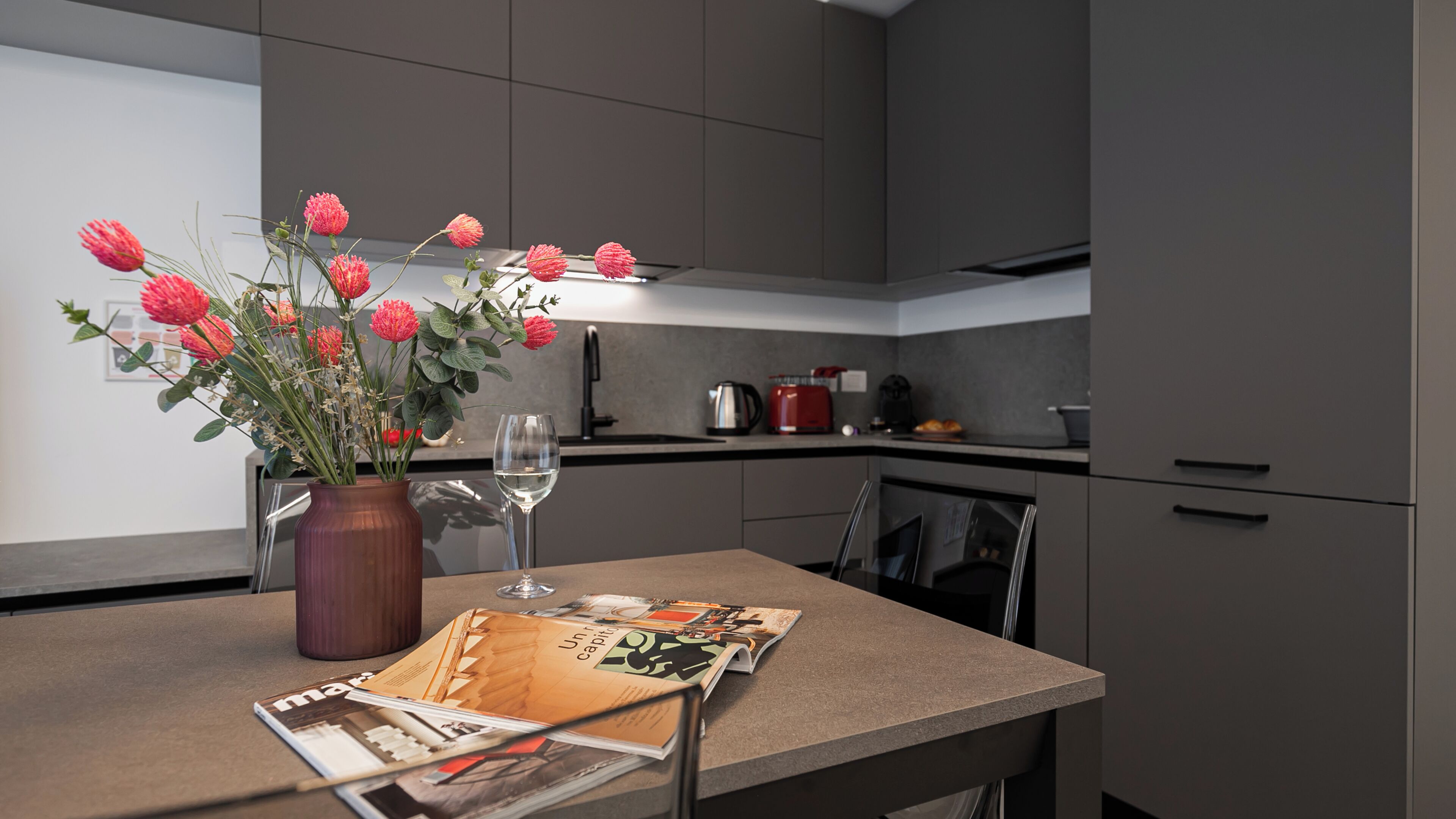 Eine moderne Küche mit grauen Schränken, einer Vase mit roten Blumen, einem Weinglas und einer Zeitschrift auf der Arbeitsfläche.
