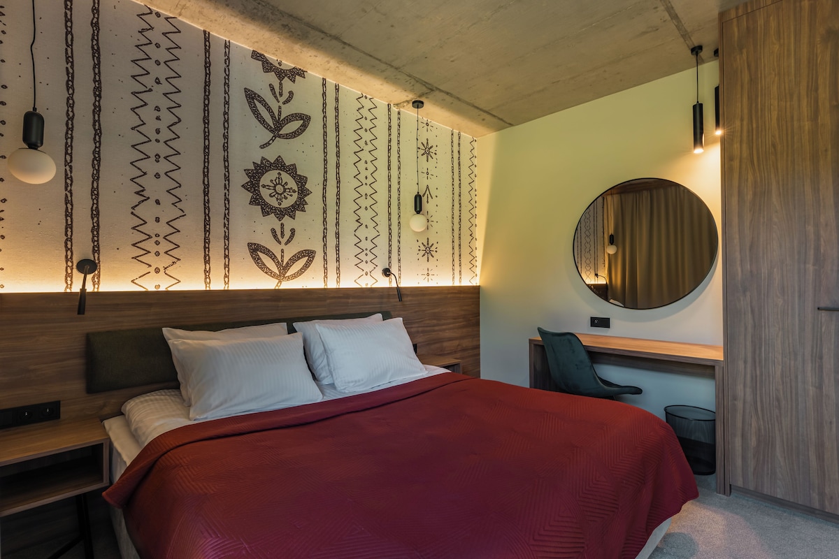 Un dormitorio moderno con cabecera de madera, cama con ropa de cama blanca y roja, un escritorio con silla y un espejo redondo en la pared.