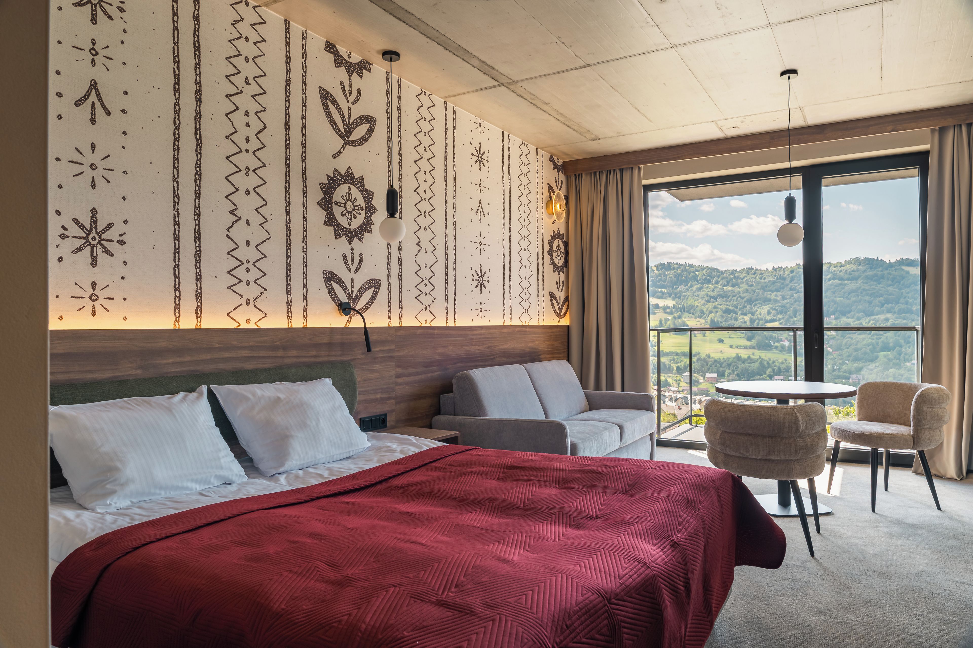 A hotel room with a large bed, a sofa, a small table, and a view of a green landscape through the window.