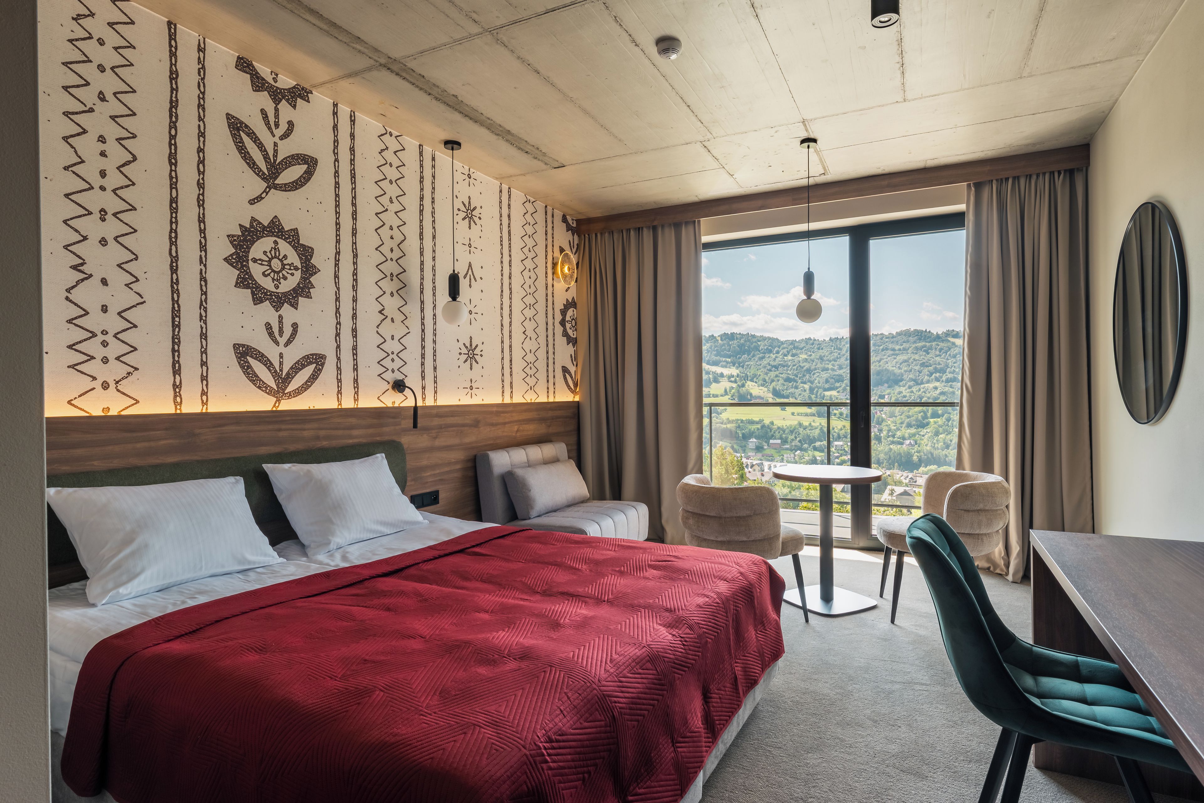 A hotel room with a large bed, two bedside tables, a desk, and a chair. There is a window with a view of a green landscape.