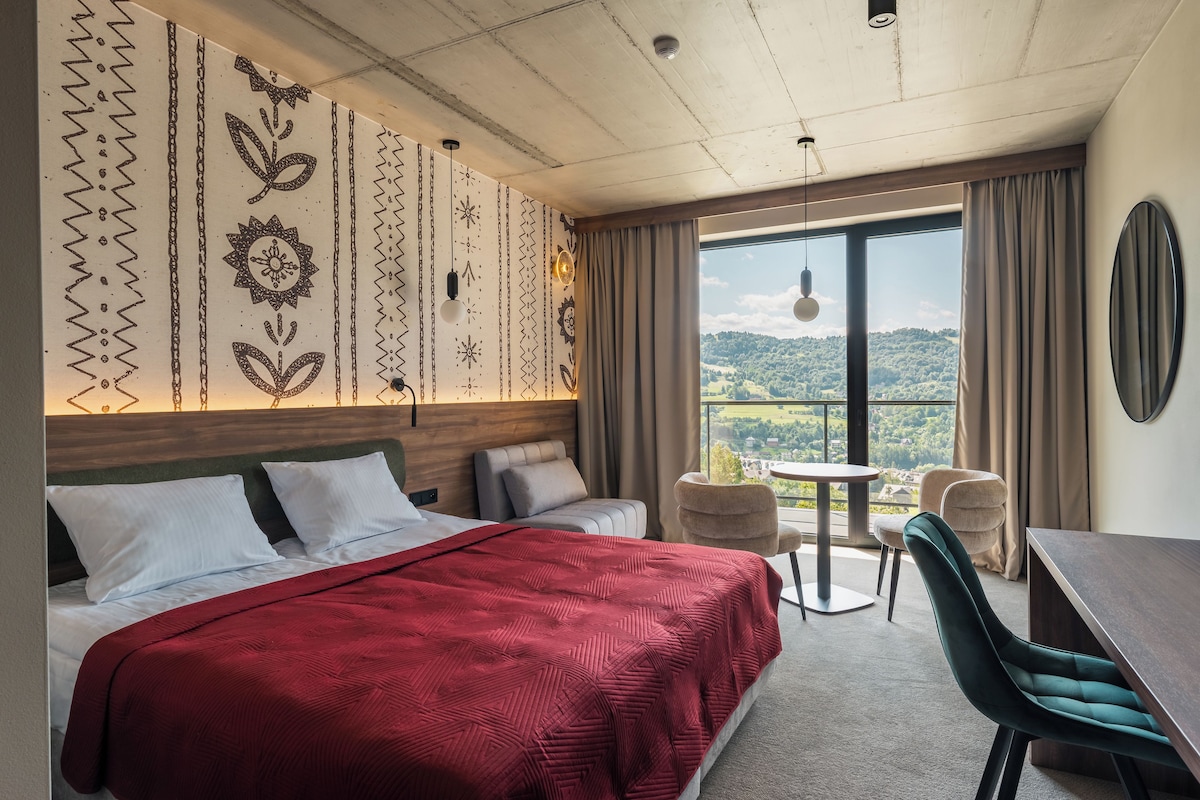 Habitación de hotel con una cama grande, dos mesitas de noche, un escritorio y una silla. Se observa un ventanal con vistas a un paisaje verde.