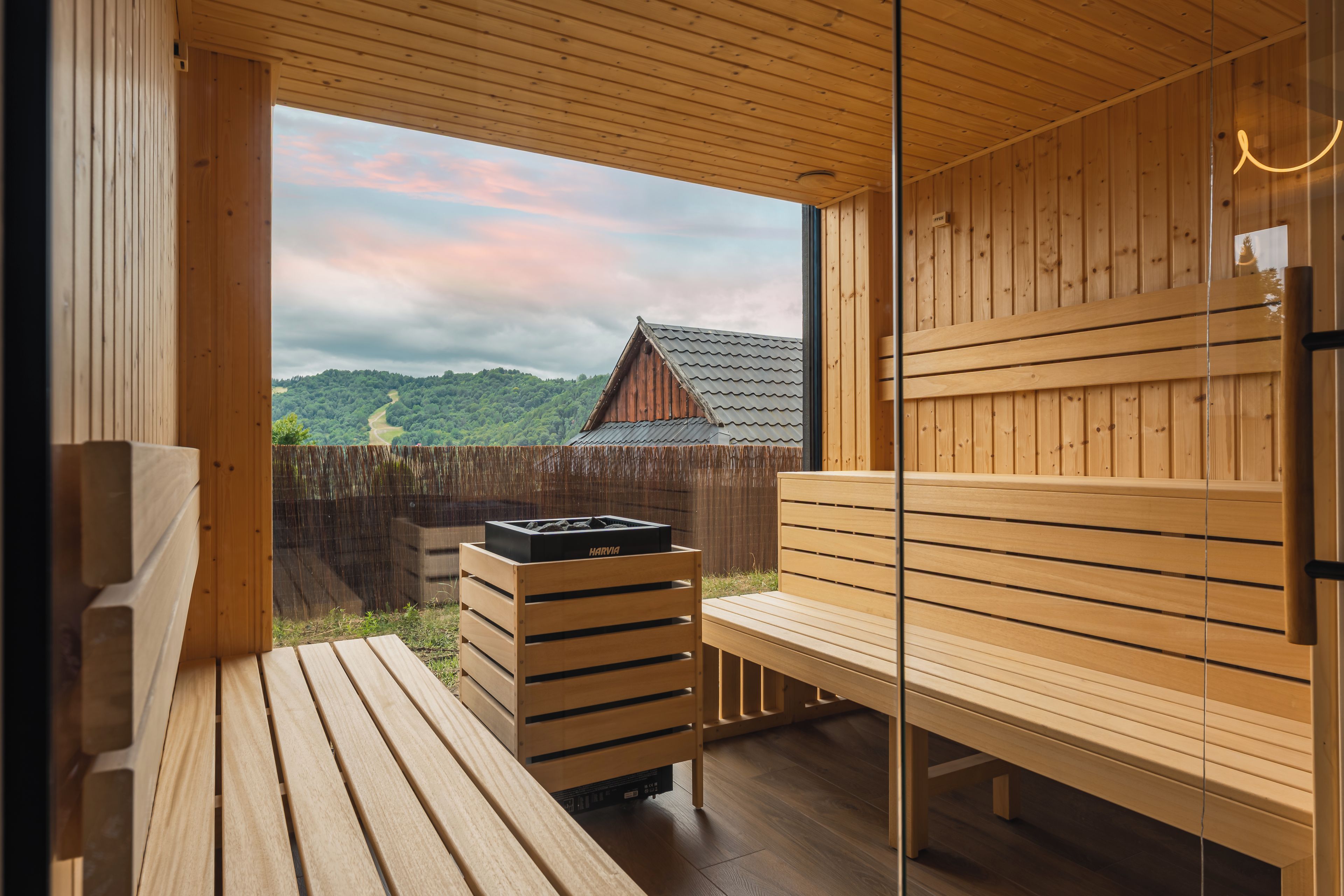 A wooden sauna with benches and a Harvia sauna heater visible through a glass door.
