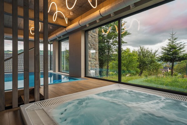 A modern indoor pool area with a hot tub, large windows, and a view of the surrounding landscape.