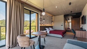 Habitación de hotel moderna con una cama grande, un escritorio, una mesa redonda y un balcón con vistas al bosque.