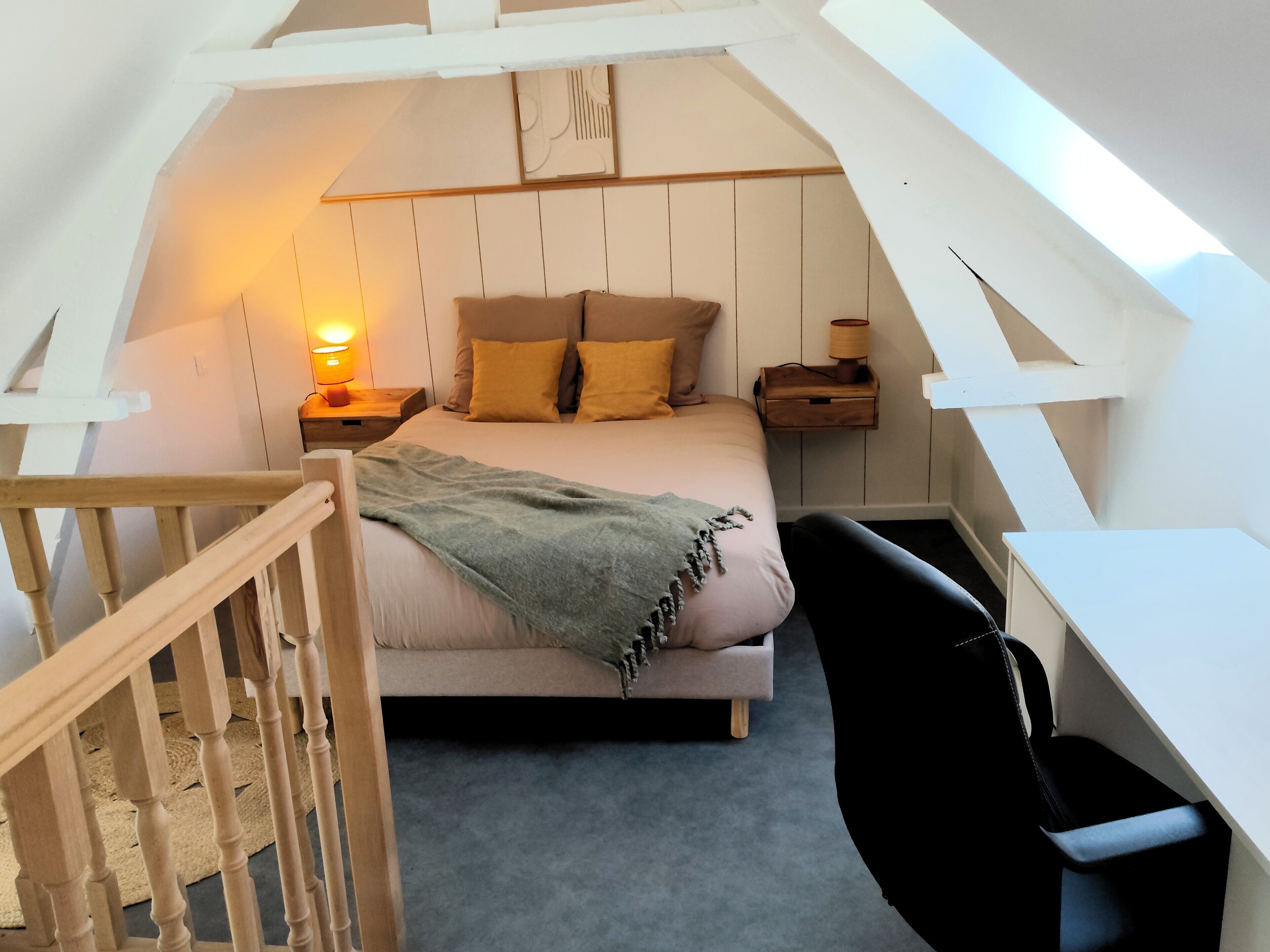 Un dormitorio con techo inclinado, una cama con almohadas, una mesita de noche con lámpara, una silla y un pequeño mueble de madera.