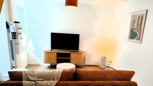 Olohuoneessa on ruskea sohva, puinen tv-taso, jossa on televisio, lattiavalaisin ja seinällä kehystetty taulu.