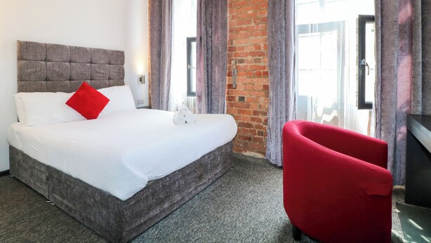 Una habitación de hotel con una cama, una silla roja, una pared de ladrillo y una ventana con cortinas.