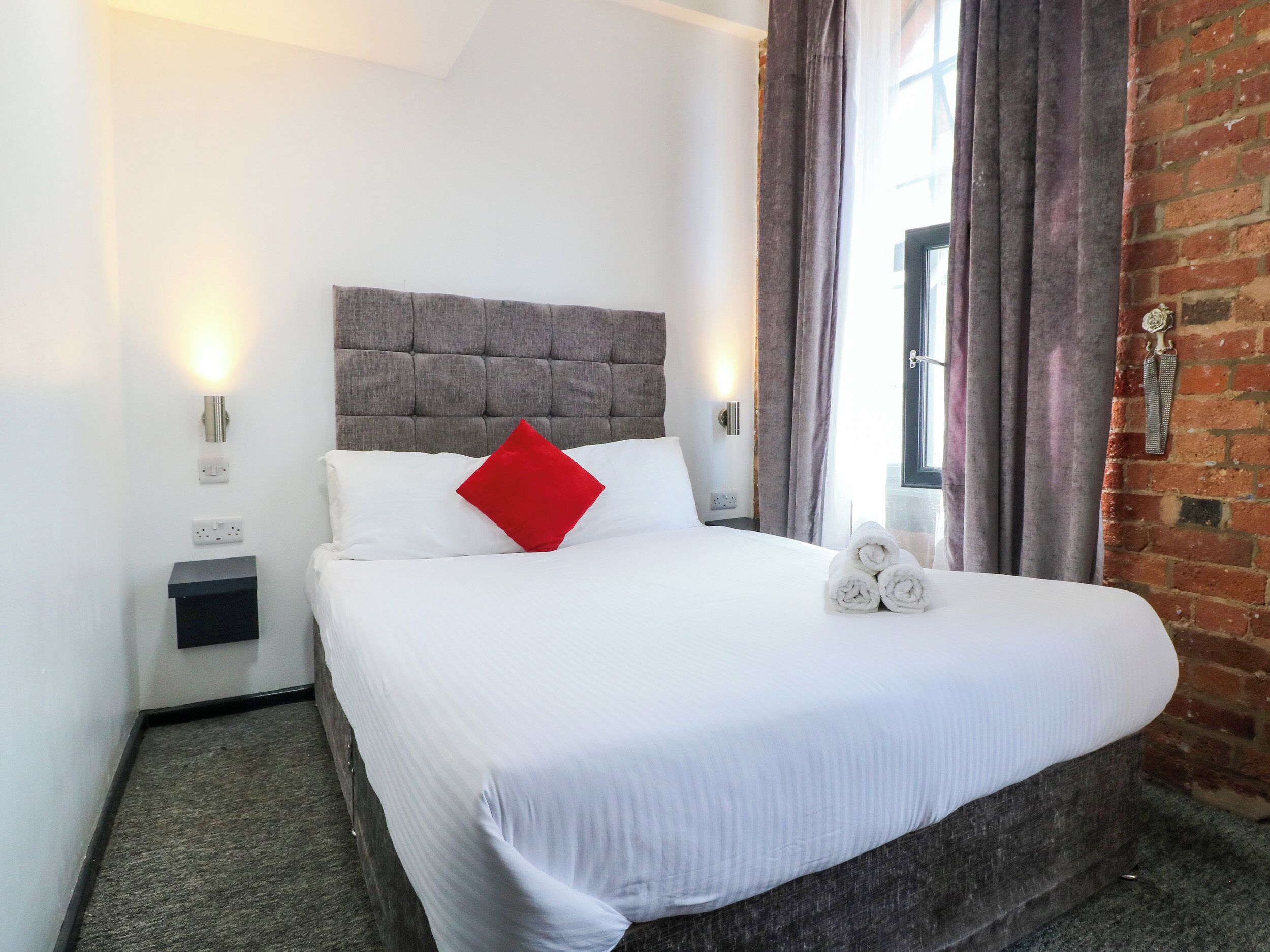 A neatly made bed with white linens, a red pillow, and rolled towels on the side.