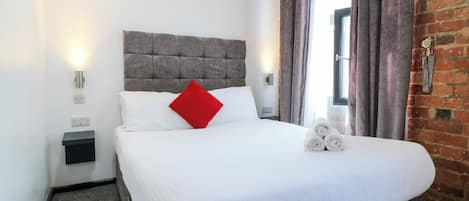 Una cama bien hecha con sábanas blancas, una almohada roja y toallas enrolladas a un lado.