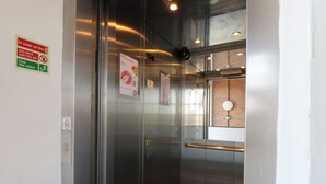 Interior de un ascensor con panel digital, instrucciones de emergencia y barandilla.