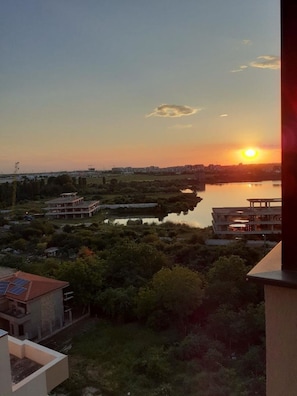 A sunset view from a balcony overlooking a body of water, with buildings and greenery in the foreground. - Cozy 1-bedroom apartment in gorgeous Constanta with AC (Constanta)