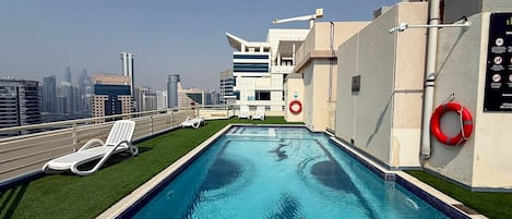 Une piscine sur le toit offrant une vue sur la ville, des chaises longues blanches et un cerceau de sauvetage.