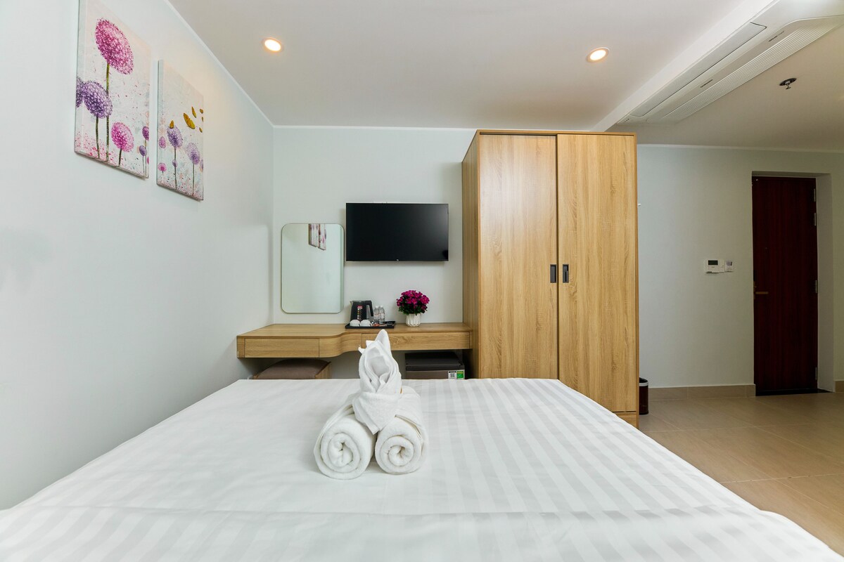 A hotel room with a bed, a wooden wardrobe, a flat-screen TV, a small table with a coffee maker, and a painting on the wall.