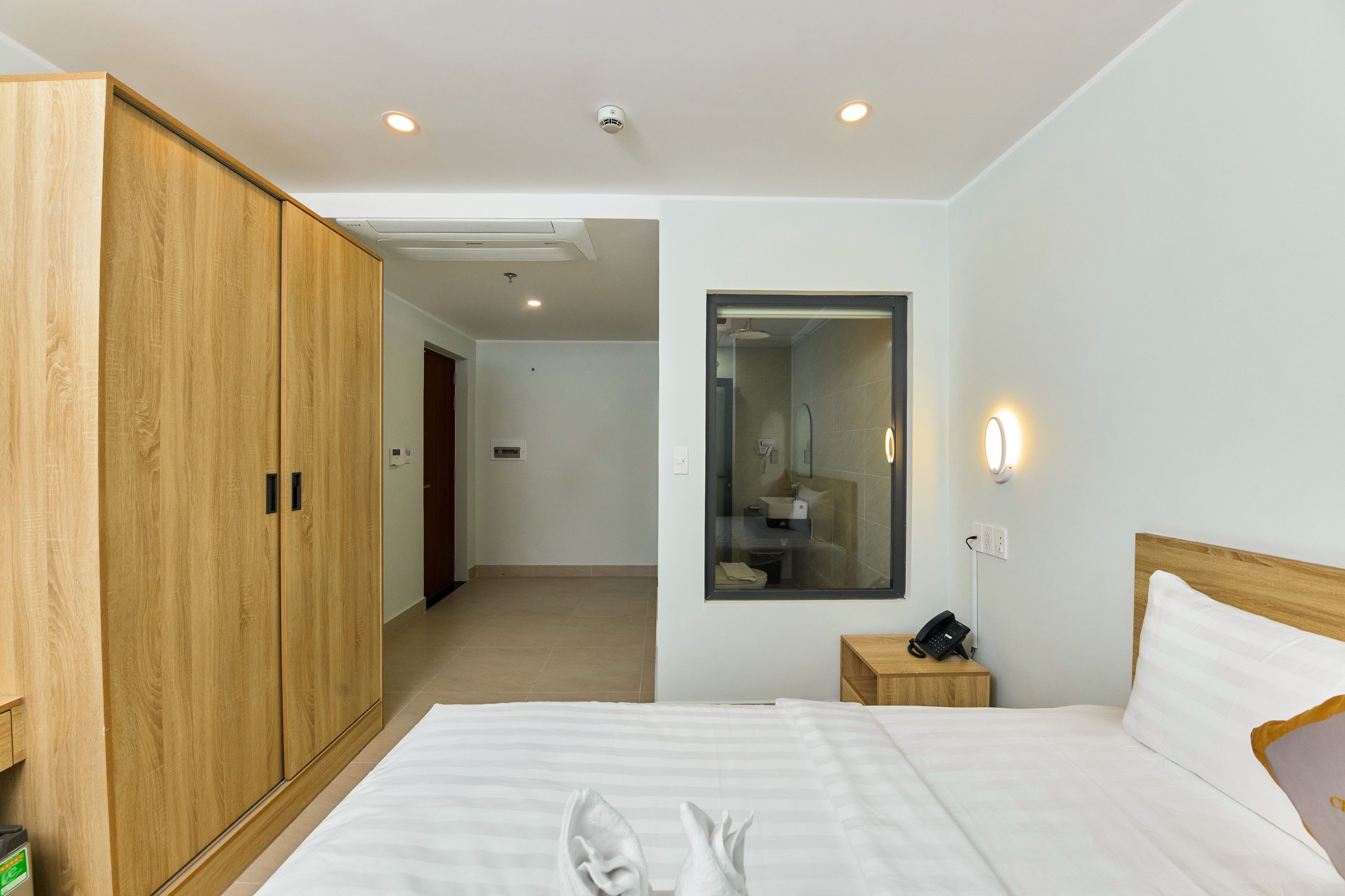 Habitación de hotel con armario de madera, cama con ropa blanca, mesita de noche con lámpara y espejo en la pared.