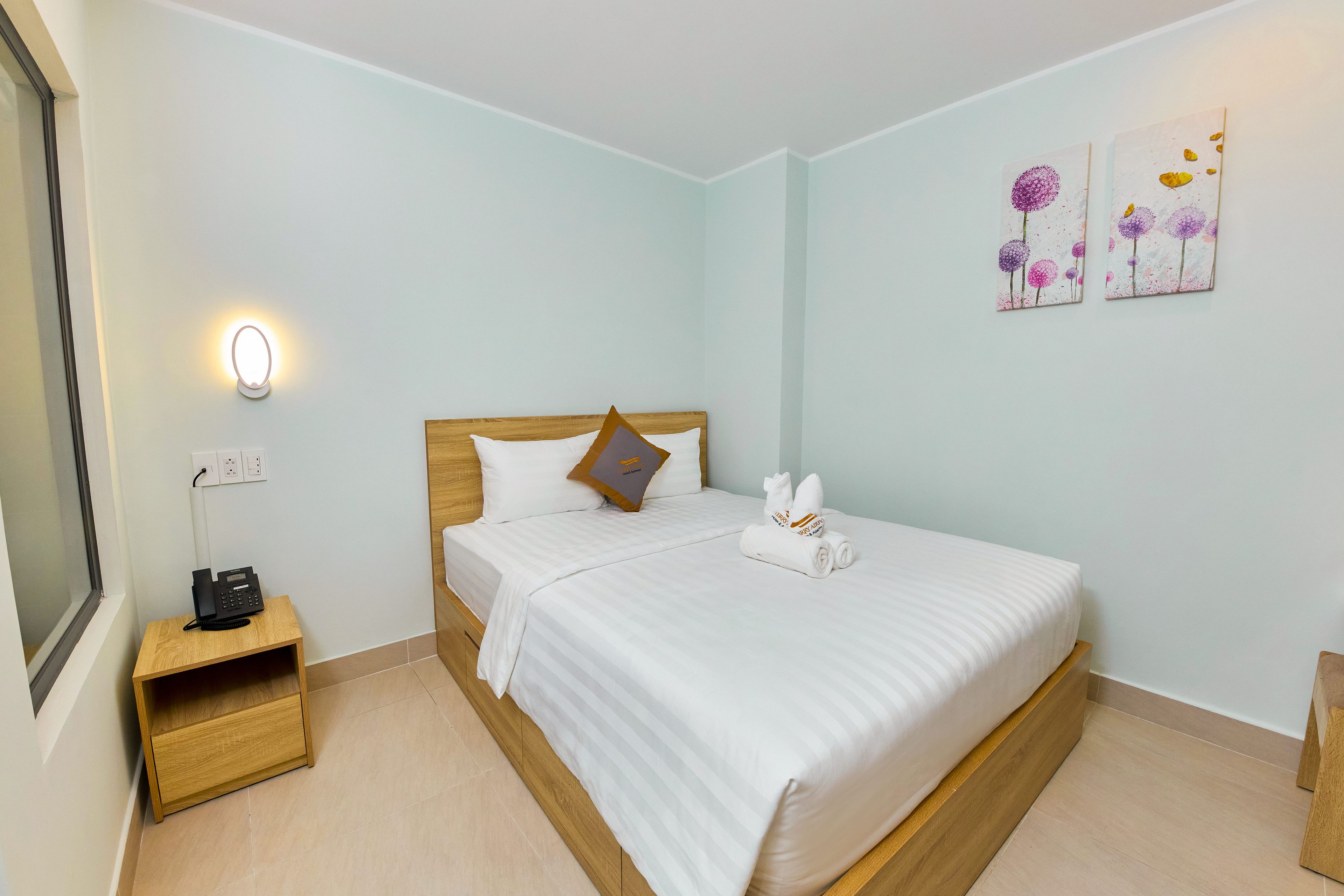 Una habitación de hotel con una cama, una mesita de noche con un teléfono, una lámpara de pared y una obra de arte.