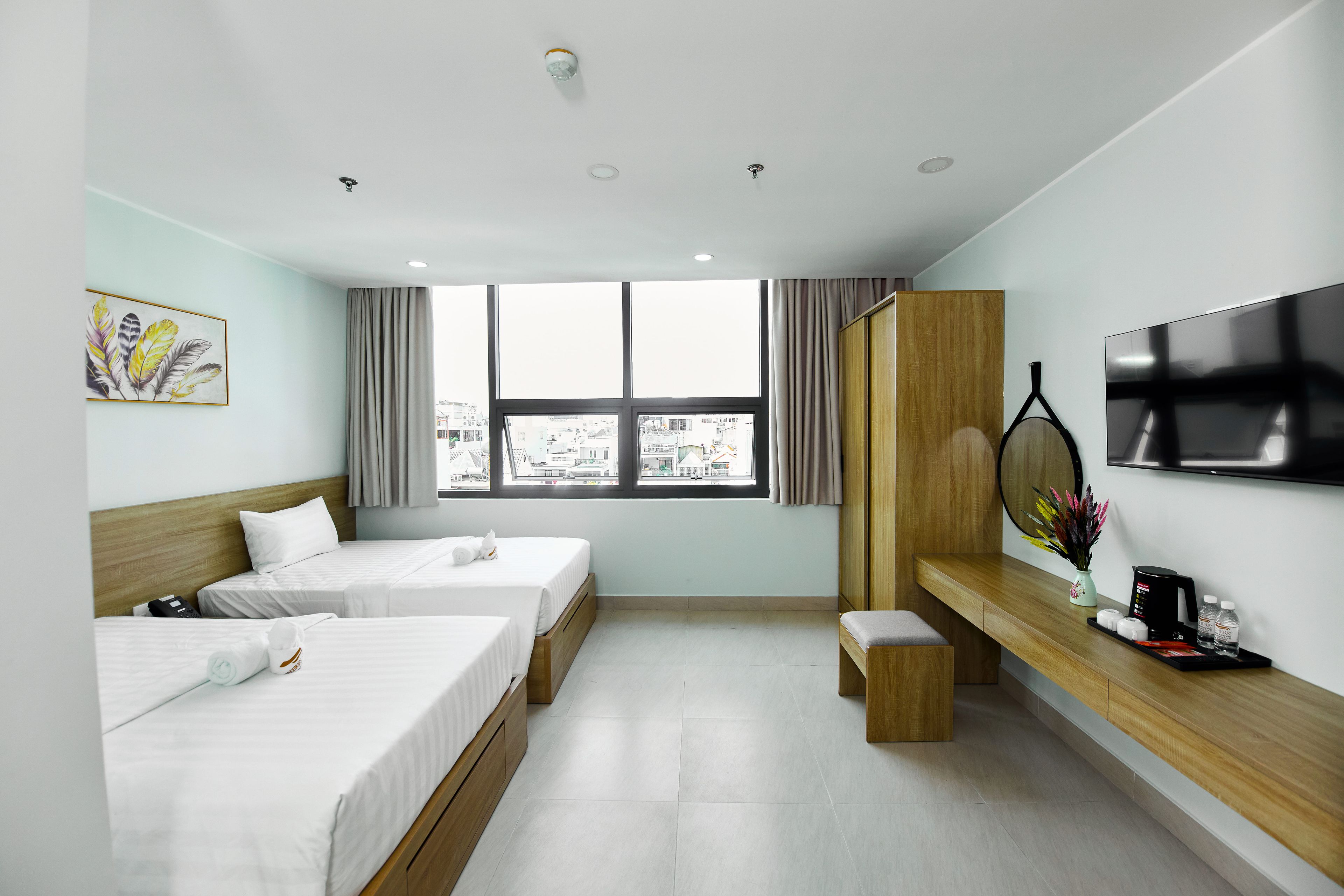 A hotel room with two beds, a TV, a window with a city view, and a wooden desk with a vase and a coffee maker.