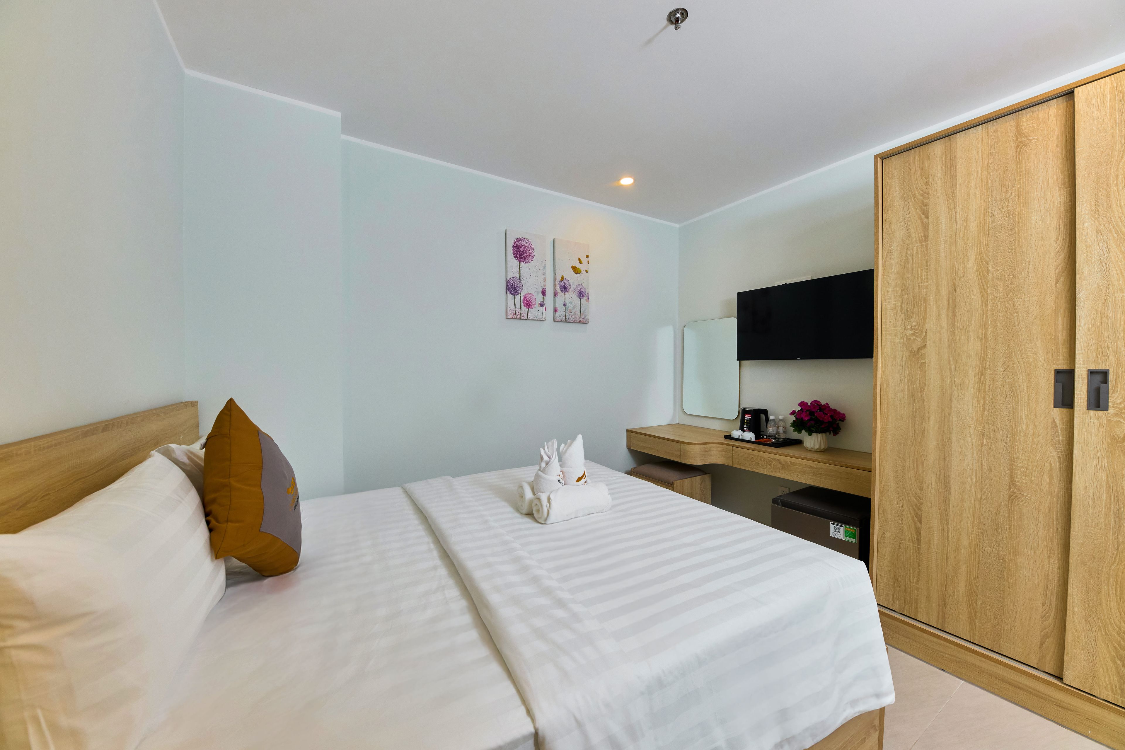 A hotel room with a bed, a wooden wardrobe, a bedside table with a mirror, and artwork on the wall.