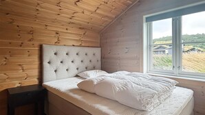 Un dormitorio con techo de madera, cabecera capitonada, cama con ropa de cama blanca y ventana con vistas a una casa y césped.