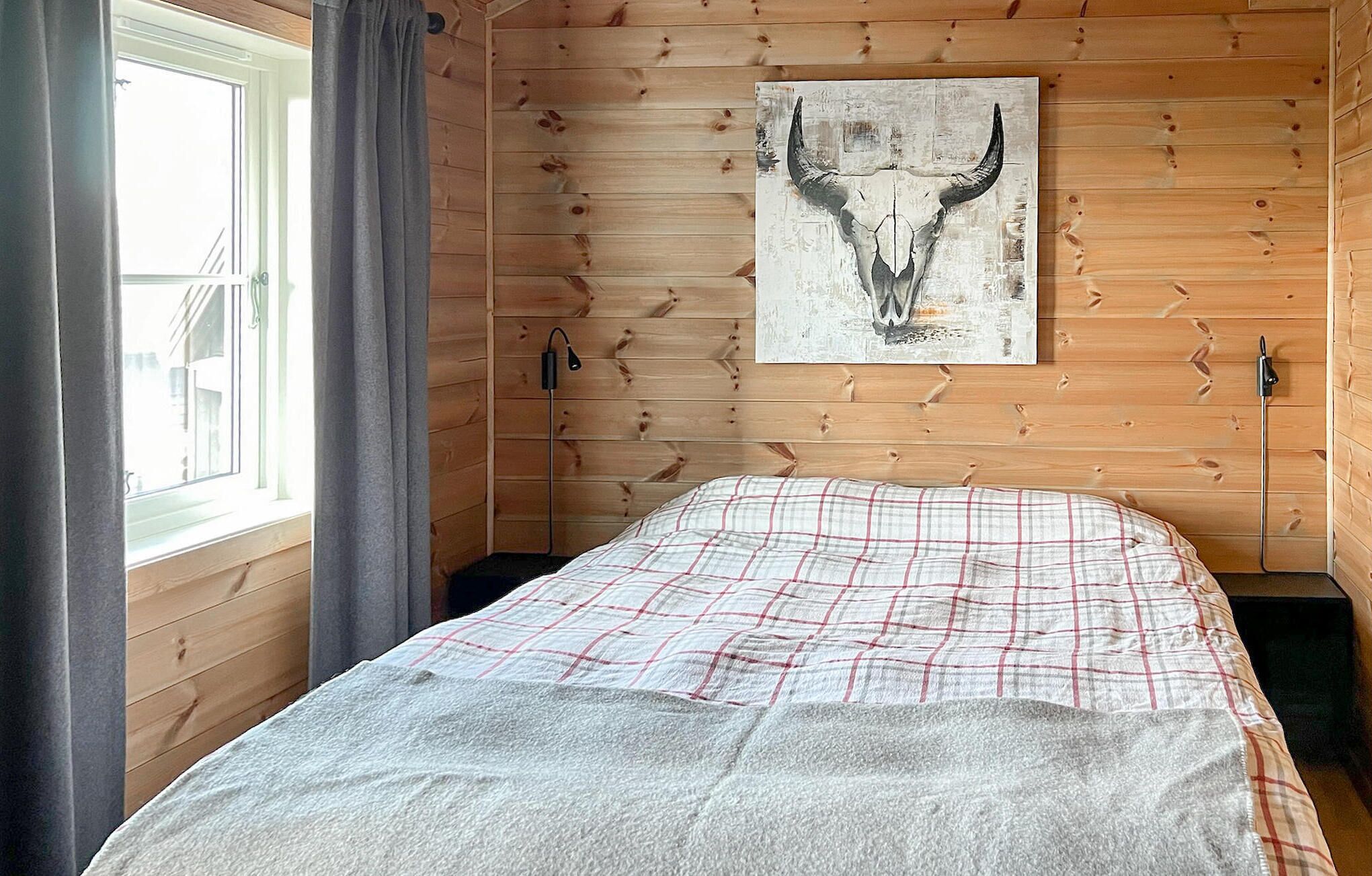 A room with wooden walls, a bed with a plaid bedspread, a mounted artwork of a bull's skull, and a window with curtains.