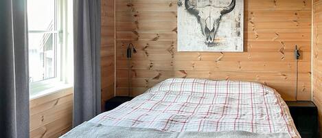 A room with wooden walls, a bed with a plaid bedspread, a mounted artwork of a bull's skull, and a window with curtains.