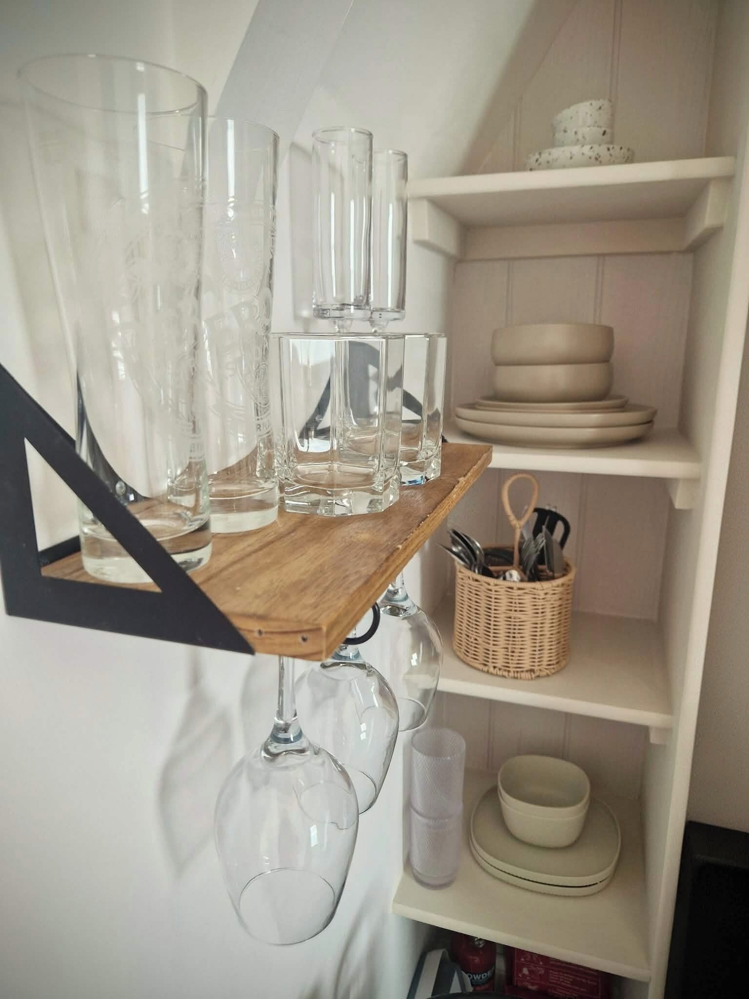 A kitchen with a wooden shelf holding various glassware and a wicker basket with utensils.