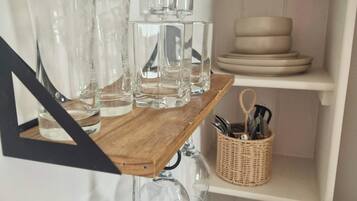 A kitchen with a wooden shelf holding various glassware and a wicker basket with utensils.