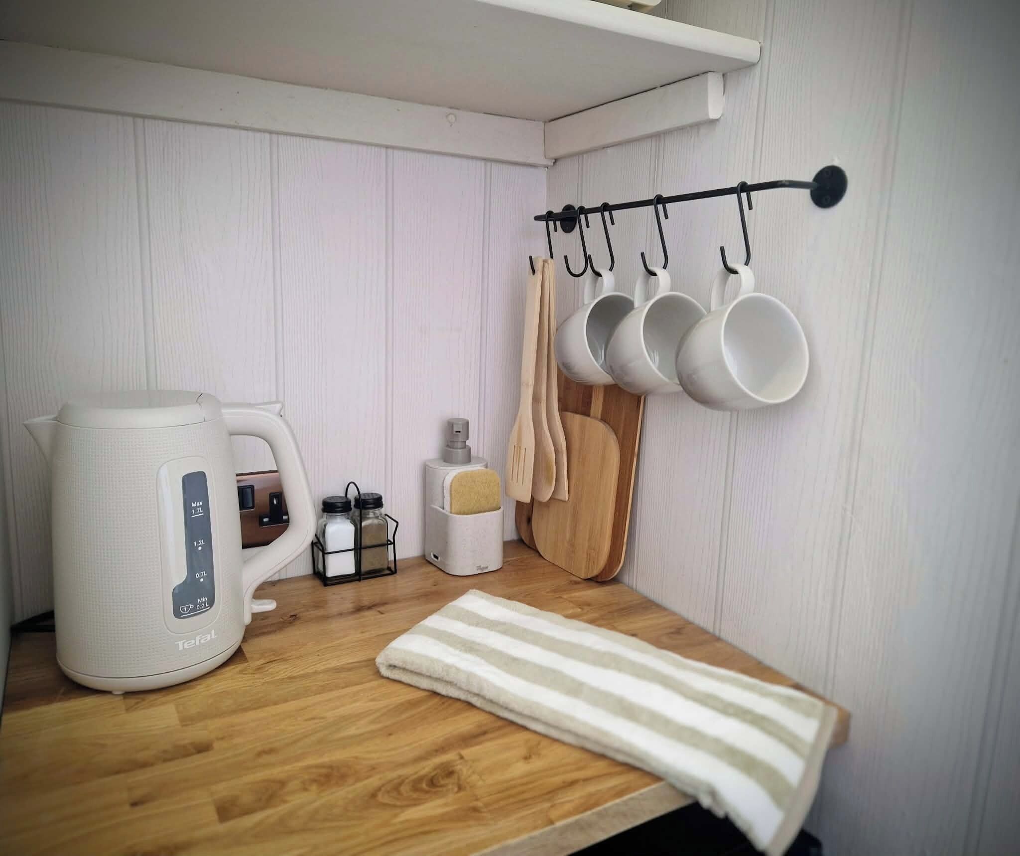 Eine Küchenzeile mit Kaffeemaschine, Wasserkocher, Gewürzregal, hängenden Tassen und einem gestreiften Handtuch auf einer Holzausziehtischplatte.