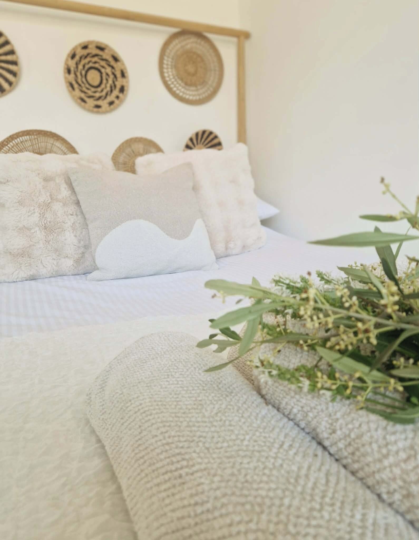 Un lit avec une couette blanche et des oreillers, une tête de lit en osier et une plante posée sur le lit.
