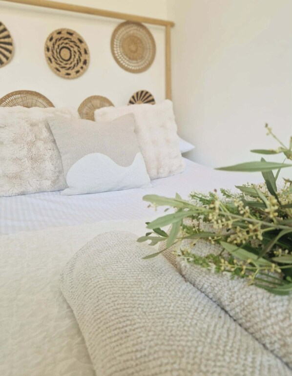 Una cama con edredón blanco y almohadas, un cabecero tejido y una planta sobre la cama.
