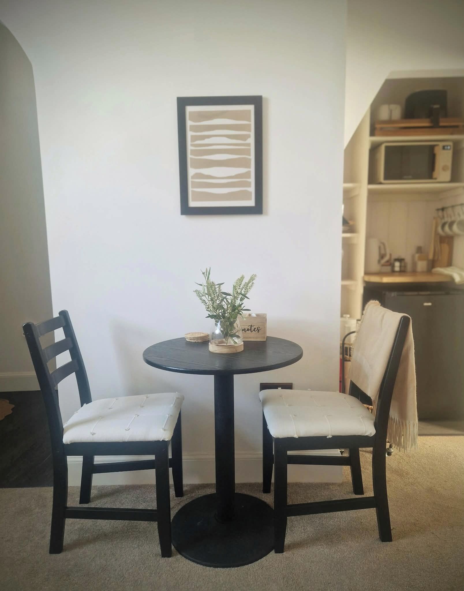 A dining area with a round table, two chairs, a vase with a plant, and a framed picture on the wall.