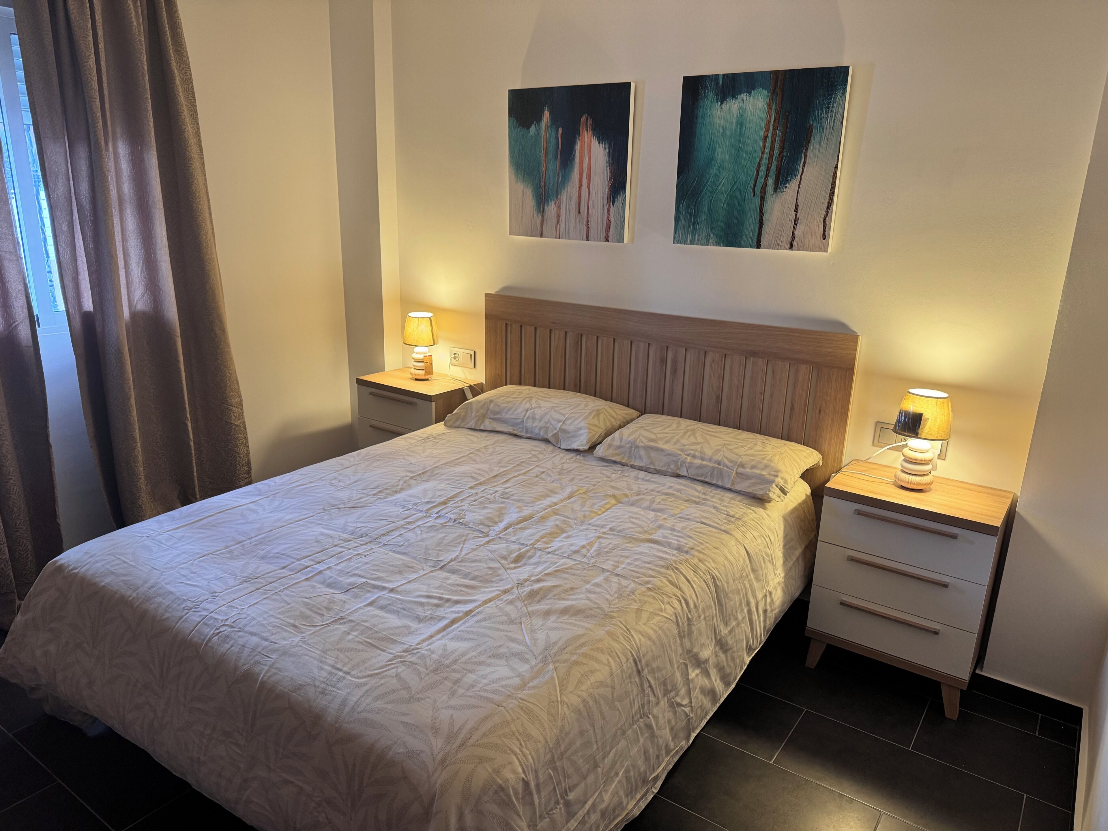 A neatly made bed with a wooden headboard, two bedside tables with lamps, and two abstract paintings on the wall.