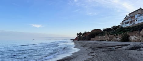 A coastal beach with a large building on a cliff, waves approaching the shore, and a clear sky.