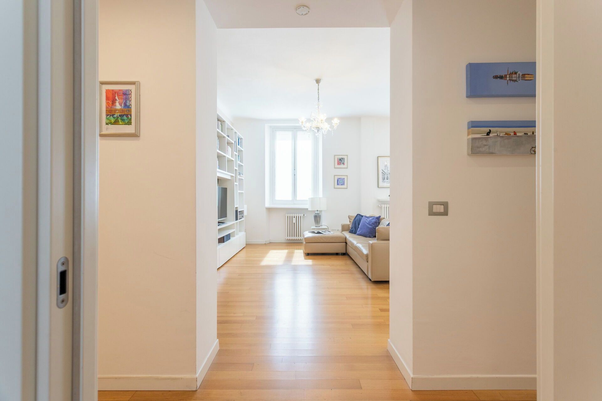 A hallway leading to a living room with a sofa, a chandelier, and a bookshelf.