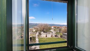 Blick durch ein Fenster auf ein Steinhaus, eine Wiese und ferne Berge.