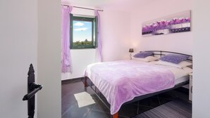 Ein Schlafzimmer mit einem Bett, einem Fenster mit Vorhängen, einem an der Wand hängenden Bild und einem Nachttisch mit einer Lampe.