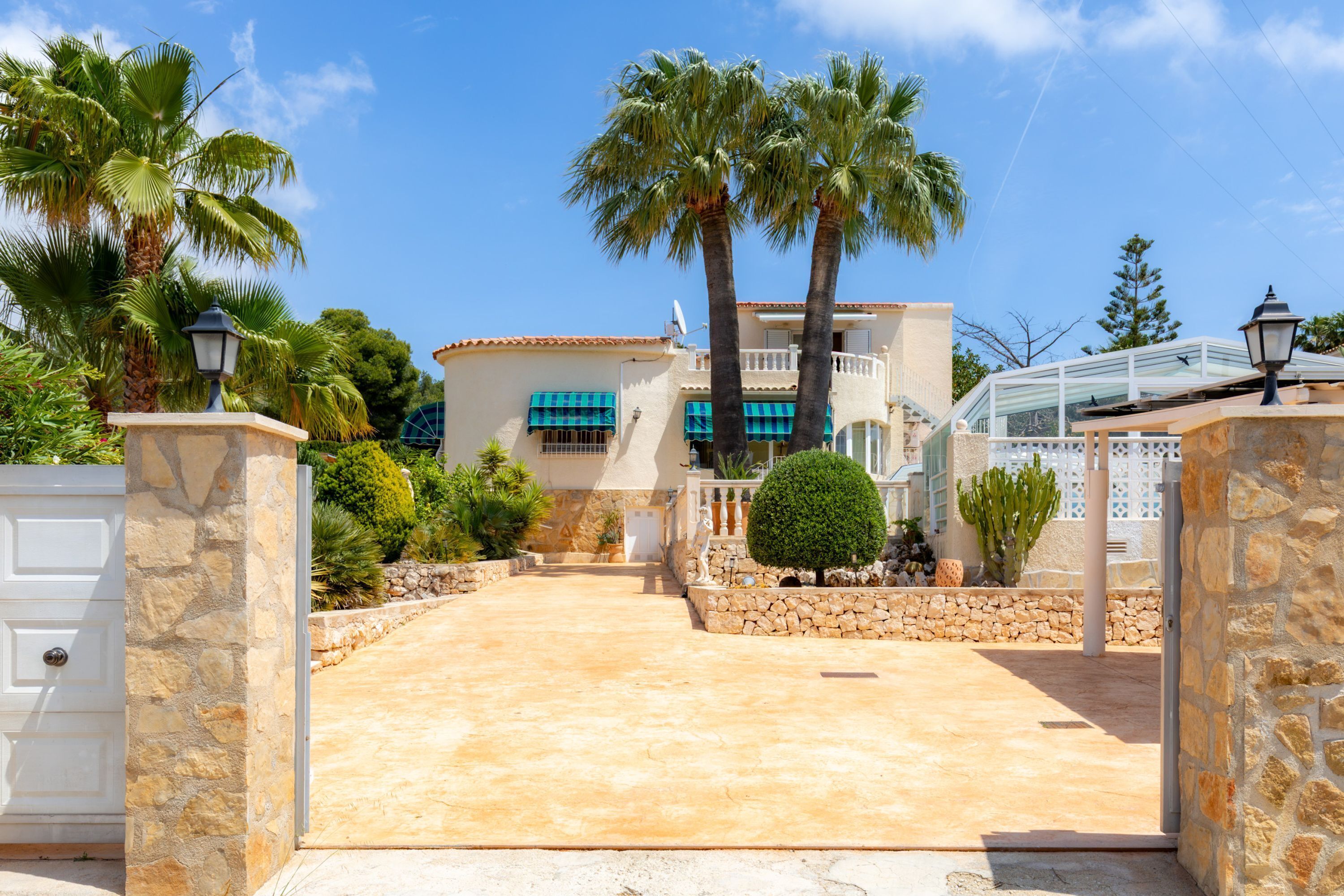 A property with a stone entrance, a paved driveway, palm trees, and a building with a tiled roof.