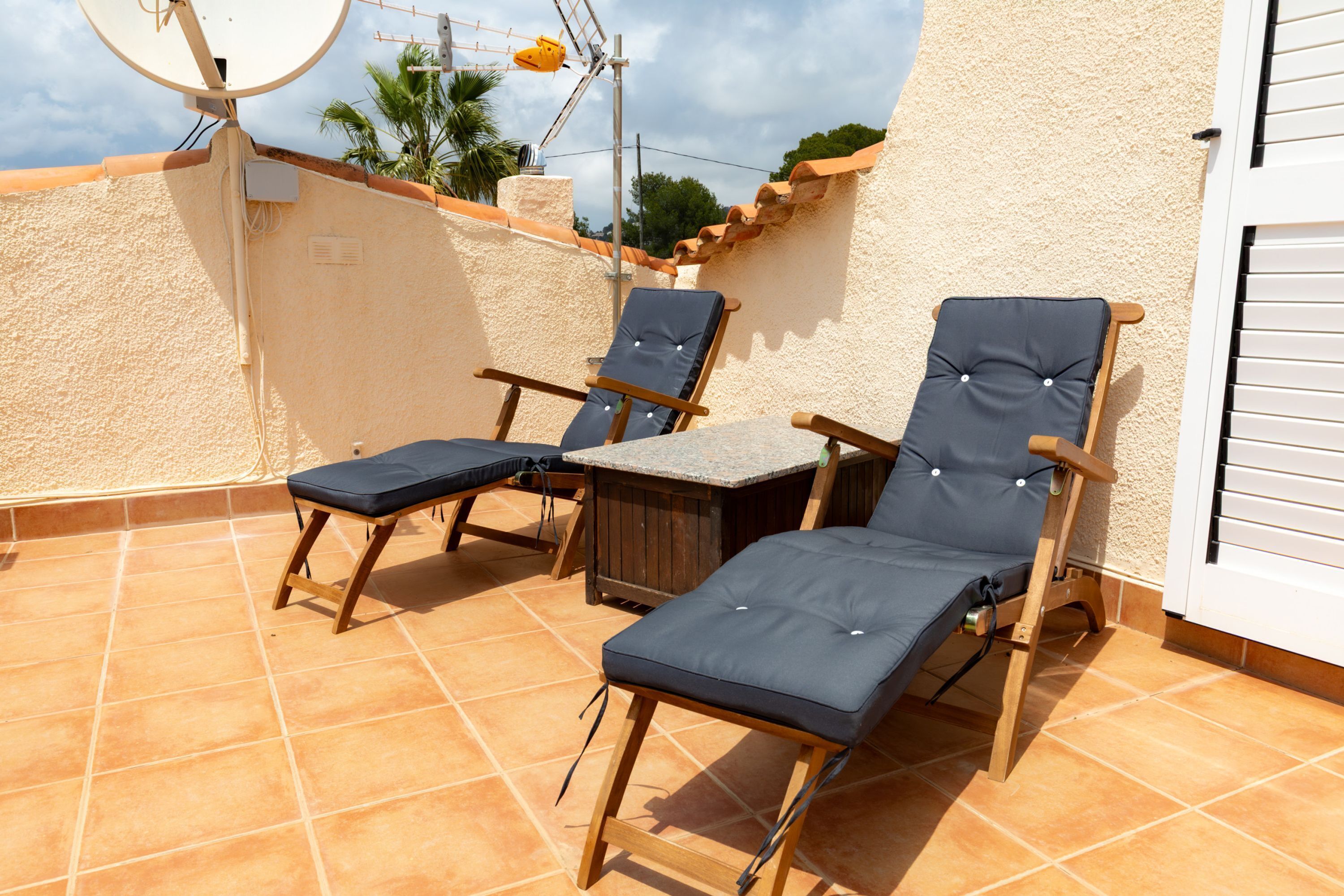 A rooftop terrace with two lounge chairs and a small table, surrounded by a tiled floor and a white wall.