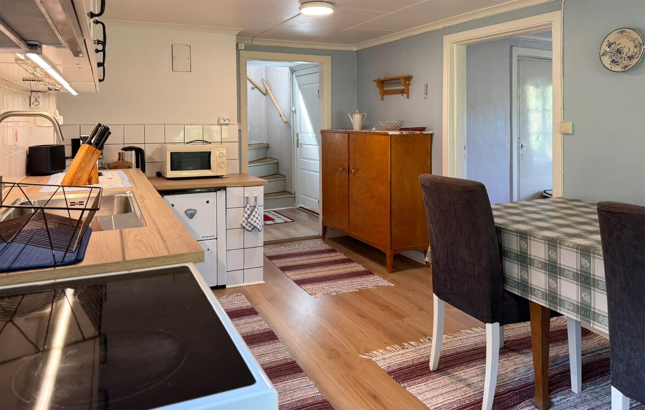 A compact kitchen with a microwave, stove, and wooden counter. There is a dining area with a checkered tablecloth and chairs.