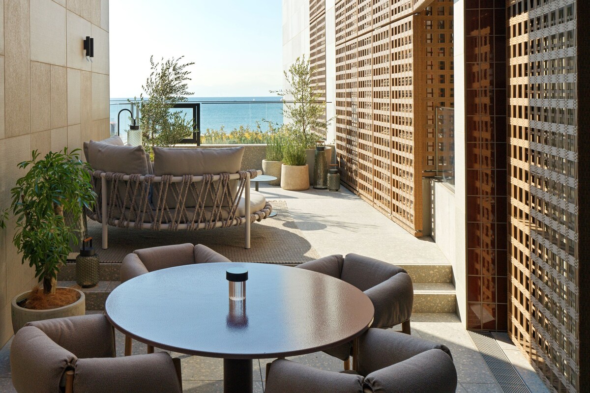 A terrace with a round table and chairs, potted plants, and a view of the sea.