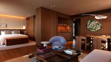 A modern hotel room with a bed, a glass coffee table, a seating area with chairs, a small table with books, and a wall-mounted flower arrangement.
