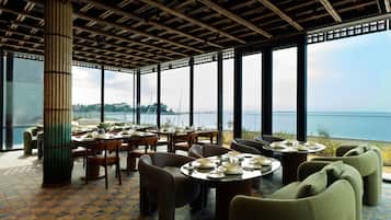 A restaurant with round tables and chairs, large windows, and a view of the sea.