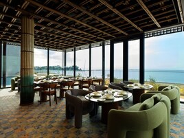 A restaurant with round tables and chairs, large windows, and a view of the sea.