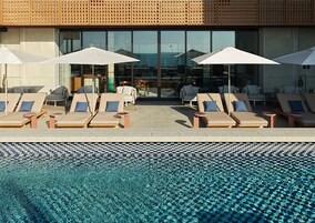 A poolside area with lounge chairs and umbrellas, a checkered pool edge, and a building with large windows in the background.