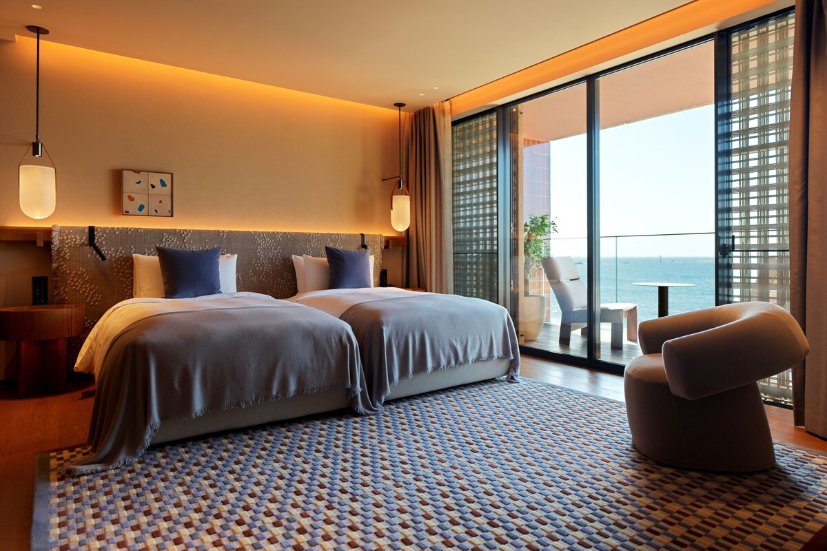 A modern hotel room with a large bed, a seating area, and a balcony with a view of the sea.