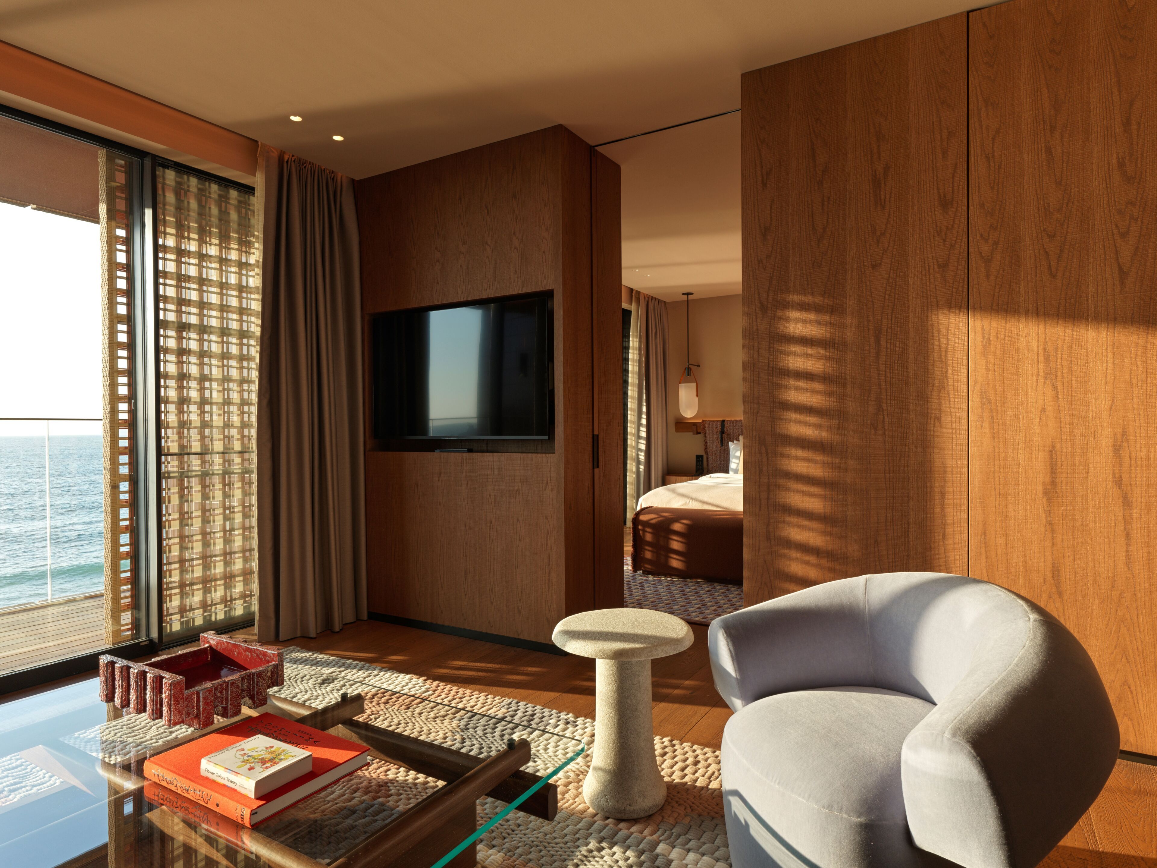 Una habitación de hotel moderna con un ventanal grande, un televisor de pantalla plana, un sillón cómodo y una pequeña mesa con libros.