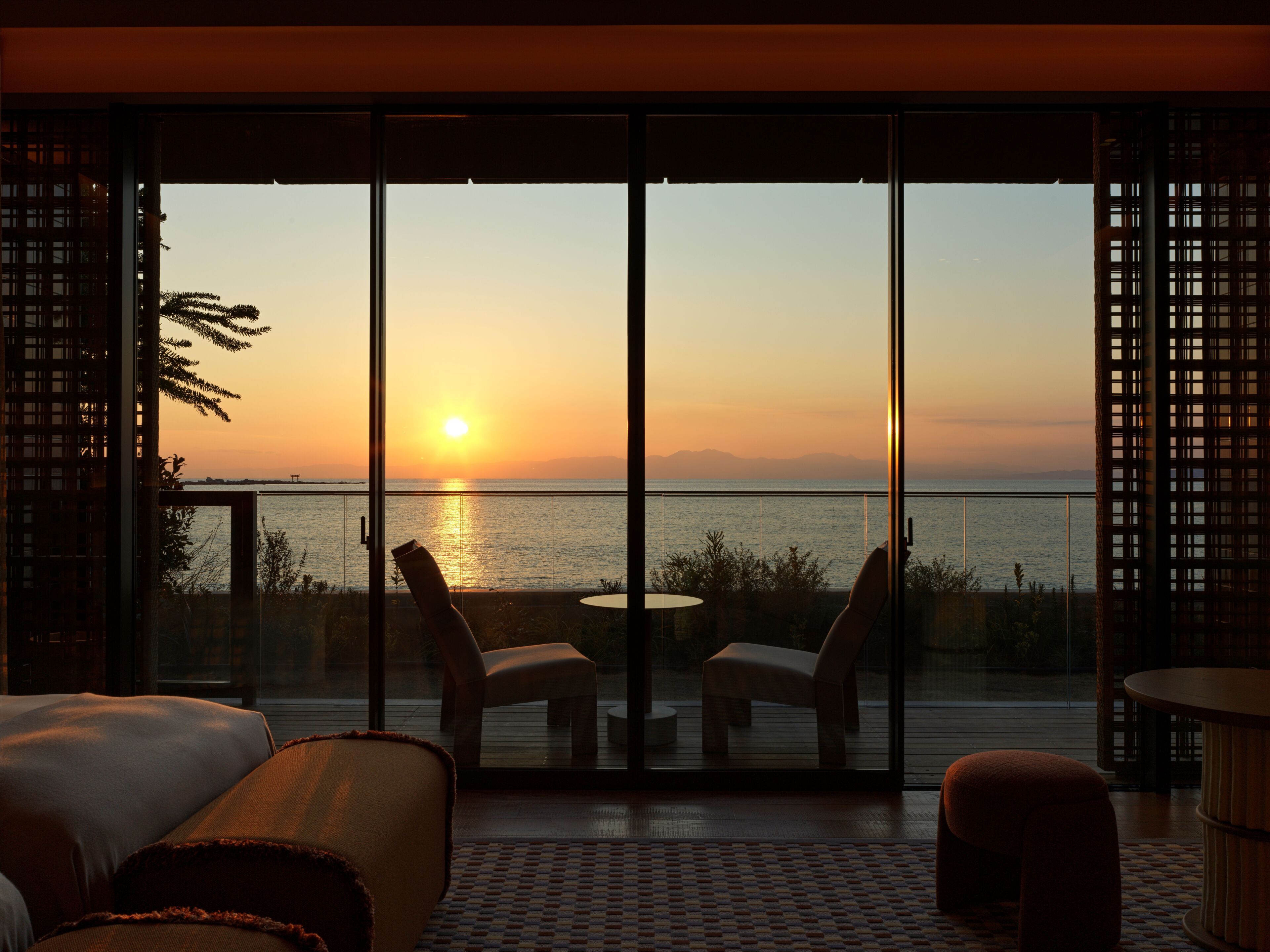 A room with a large sliding glass door leading to a balcony overlooking a sunset over water.