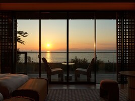 A room with a large sliding glass door leading to a balcony overlooking a sunset over water.