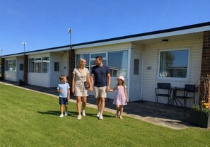Uma família caminhando em frente a uma casa móvel com garagem, um pátio com cadeiras e um pequeno jardim.