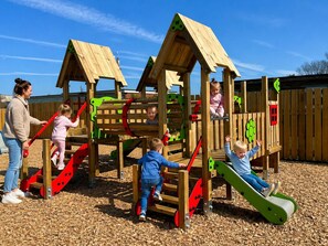 A wooden play structure with slides and climbing elements, surrounded by a wooden fence. - Comfortable & homely 2-bed Chalet with FREE passes to swimming pool, gym & club (GREAT YARMOUTH)