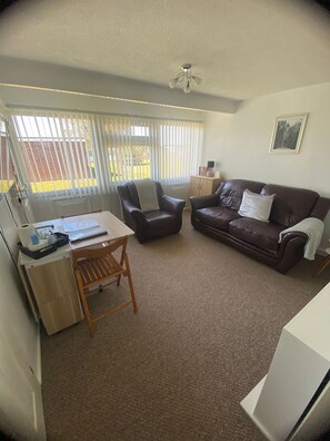 A living room with a brown leather sofa, a wooden chair, a desk with a computer, and a window with blinds. - Comfortable & homely 2-bed Chalet with FREE passes to swimming pool, gym & club (GREAT YARMOUTH)